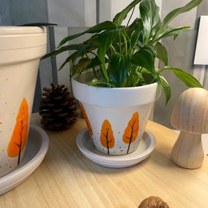 Peut inclure: Un pot de fleurs en céramique blanche avec sa soucoupe assortie, orné d'arbres orange peints à la main avec des troncs bruns et de petits points orange. Une plante verte est installée à l'intérieur. Une pomme de pin, un champignon en bois et deux noix.