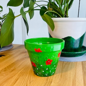 Peut inclure: Un pot de fleurs en terre cuite vert avec des fleurs rouges et des points blancs peints dessus. Le pot est posé sur une surface en bois.