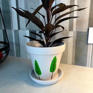 Peut inclure: Un pot de fleurs en céramique blanche avec sa soucoupe assortie, orné d'arbres peints à la main en vert et de petits points jaunes. Une plante aux feuilles sombres et pointues pousse au centre du pot, qui présente un rebord large.