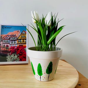 Peut inclure: Un pot de fleurs blanc orné de motifs d'arbres peints à la main en vert avec de petits points jaunes et rouges, contenant des fleurs blanches ressemblant à des tulipes aux longues feuilles vertes. Un cadre représentant Colmar est visible.