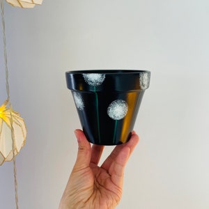 Peut inclure: Pot de fleurs en terre cuite noire avec des motifs de pissenlits blancs et des tiges vertes.