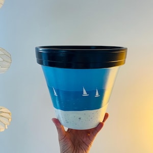 Peut inclure: Un pot en céramique bleu et blanc avec un bord noir, peint avec une scène de paysage marin de voiliers sur l'océan et une plage de sable.