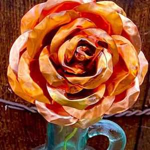 May include: A copper rose with a textured, hammered finish, displayed in a clear glass vase with a green stem.
