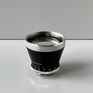 Peut inclure: Un objectif d'appareil photo noir et argenté avec le texte "Pro-Tessar" et des marquages de mesure. L'objectif a une bague argentée striée en haut et en bas, avec un corps noir. L'objectif est fabriqué en Allemagne.