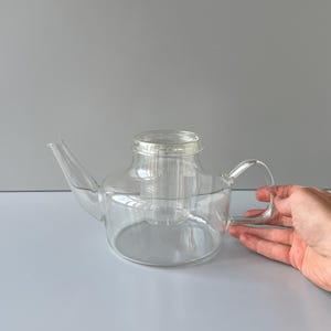 May include: A transparent glass teapot with an integrated infuser. The teapot features a curved spout and a handle. The image shows the teapot on a white surface with a grey background.