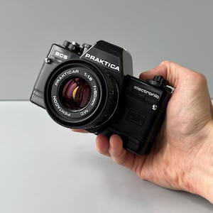 May include: Black Praktica camera with a Pentacon lens. The lens has the text "PRAKTICAR 1:1.8 f=50mm MC". The camera has the text "PRAKTICA" and "electronic". The camera is held in a hand.