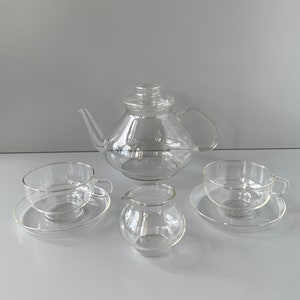 Könnte beinhalten: Eine klare Glaskanne mit Deckel, zwei klare Gläser mit Untertassen und ein klarer Glaskännchen. Das Set befindet sich auf einem weißen Hintergrund.