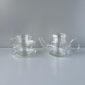 May include: A clear glass tea set, including a teapot with a handle and spout, and two teacups with saucers. The set is made of transparent glass, allowing the contents to be visible. The teapot has a built-in infuser.