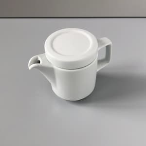 Puede incluir: Una pequeña cremera de cerámica blanca con tapa a juego. La cremera tiene un asa y un pico para verter. La tapa se asienta sobre la cremera, cubriendo la abertura. La cremera está diseñada con una estética minimalista.