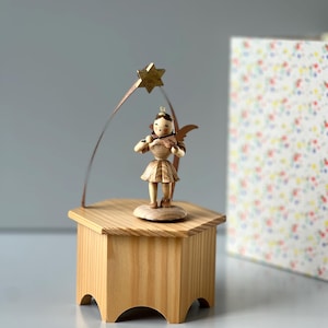 Könnte beinhalten: Eine Holzspieluhr mit einem geschnitzten Engel, der Geige spielt. Die Engelsfigur steht auf einem sechseckigen Holzsockel mit einem goldenen Stern darüber. Die Box ist aus hellem Holz gefertigt und hat ein schlichtes, elegantes Design.