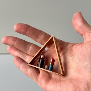 Könnte beinhalten: Ein hölzerner Dreiecksrahmen mit handbemalten Miniaturfiguren. Die Figuren stellen eine religiöse Szene dar, mit einer Figur in Schwarz, einer in Rot und Blau und einer kleinen Figur in Weiß und Gold. Der Rahmen wird in einer Hand gehalten.