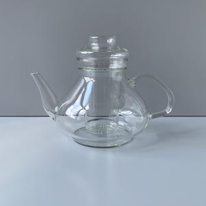 May include: A clear glass teapot featuring a wide, rounded base, a curved handle, and a slender spout. The teapot includes a removable glass infuser basket positioned in the centre and a matching clear glass lid that sits securely on the top opening.
