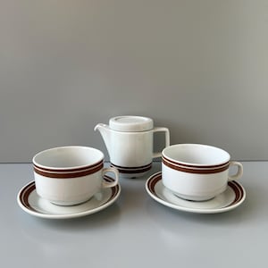 Peut inclure: Service à thé en céramique blanche avec des rayures marron. L'ensemble comprend une théière avec un couvercle, deux tasses et deux soucoupes. Les tasses et les soucoupes ont une rayure marron autour du bord. La théière a une rayure marron autour de la base.