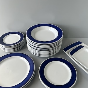 Pode incluir: Um conjunto de louça de cerâmica azul e branca, incluindo pratos, tigelas e um prato de servir. Os pratos têm uma faixa azul larga em torno da borda.