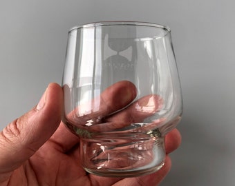 May include: A clear, stemless glass with a rounded bowl and a thick base. The glass is held in a hand and features a logo with a bow tie design and the word "sommeliers" etched on the side. The background is a solid gray.