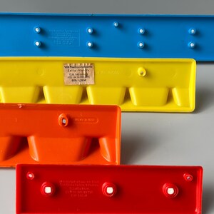 May include: Four plastic building blocks in different colours: blue, yellow, orange, and red. The blocks have a series of small holes on the top and bottom. The blue block has a label with the text "VEB Plasthousho/twaren Zella-Mais ELN 145 6 HSL 64 16 900 0201 EVP: 1,70 M Made in FVW 145 M Nr 14005".