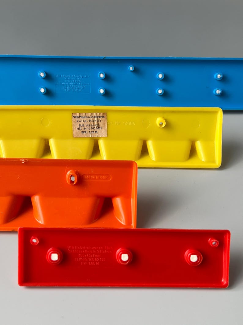 May include: Four plastic building blocks in different colours: blue, yellow, orange, and red. The blocks have a series of small holes on the top and bottom. The blue block has a label with the text "VEB Plasthousho/twaren Zella-Mais ELN 145 6 HSL 64 16 900 0201 EVP: 1,70 M Made in FVW 145 M Nr 14005".