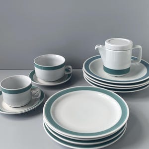 May include: A set of white teacups and saucers with a teal band around the rim. The set includes a teapot with a lid and a stack of four plates.