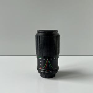 May include: A black camera lens with a ribbed grip and measurement markings. The lens has a series of numbers and colored lines indicating focal lengths and settings. The lens is cylindrical and stands upright.