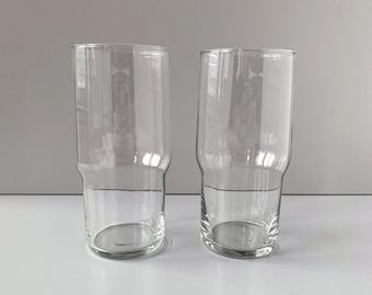 May include: Two clear, cylindrical drinking glasses with a slightly wider base and a subtle step near the bottom. The glasses are empty and appear to be made of transparent glass. The background is a plain, light gray. These glasses are suitable for water, juice, or other beverages.