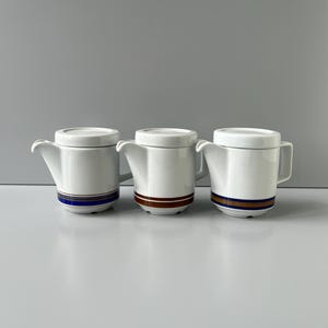 Peut inclure: Trois théières en céramique blanche avec couvercles assortis sont présentées. Chaque théière a une anse et un bec verseur. Une bande horizontale de couleur, dont le bleu, le marron et l'or, décore la base de chaque théière. Les théières sont disposées sur une surface gris clair.