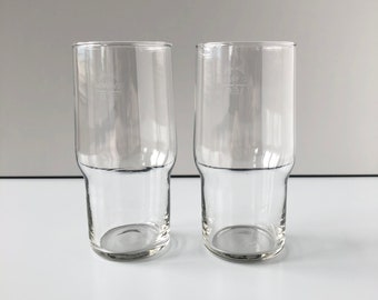 May include: Two clear glass drinking glasses with a slightly flared lip and a thicker base. The glasses are empty and sitting on a white surface.