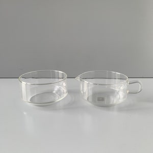 May include: Two clear glass containers, one a bowl and the other a jug with a handle. Both are empty and sitting on a white surface.