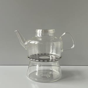 May include: A clear glass teapot with a curved handle and spout. The teapot sits on a metal base with a circular design. The design is simple and modern, suitable for brewing tea.