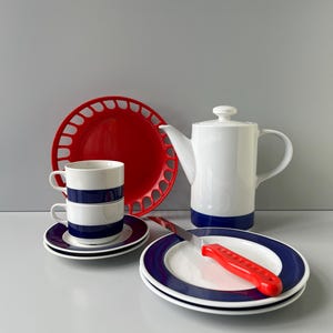May include: A white ceramic teapot with a blue base, two stacked white cups with blue stripes on matching saucers, a red decorative plate with a cutout rim, and two white plates with blue rims featuring a knife with a red handle.