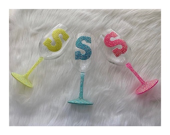 May include: Three wine glasses with a single letter 'S' on each. The glasses are decorated with rhinestones and have different colored stems: yellow, turquoise, and pink.