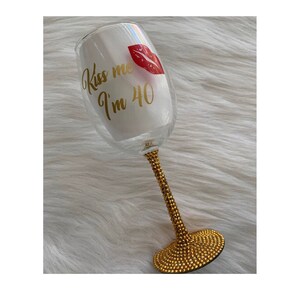 May include: A clear glass wine glass with a gold glitter stem and base. The glass has a red lip print and the text "Kiss me I'm 40" in gold lettering.