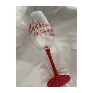 May include: A clear wine glass with a red stem and base covered in rhinestones. The glass has a personalized inscription that reads "Andrea Est. July 18, 1987 Ruby."