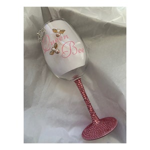 May include: A pink wine glass with a clear stem and base. The glass is decorated with rhinestones and features the text "Queen Bee" in pink lettering with a bee illustration.
