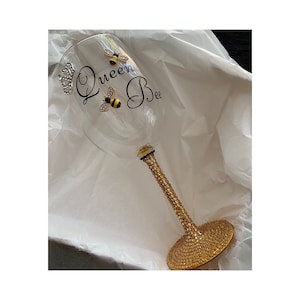 May include: A gold-rimmed glass with a gold glitter stem and base. The glass has a black and yellow bee design and the text "Queen Bee" on the side.