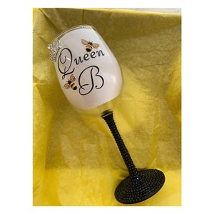 May include: A clear glass wine glass with a black stem and base. The glass is decorated with rhinestones and has the text "Queen B" with two bee illustrations.