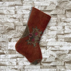 May include: A handmade Christmas stocking in shades of rust and olive green, adorned with a floral design. The stocking is made of a textured fabric and hangs against a light-colored brick wall, perfect for holiday decor.