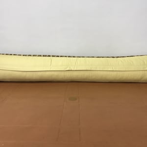 May include: A long, rectangular pillow with a light yellow back and a patterned top. The top features a mix of brown, green, and beige colors. The pillow is resting on a brown surface against a white background.