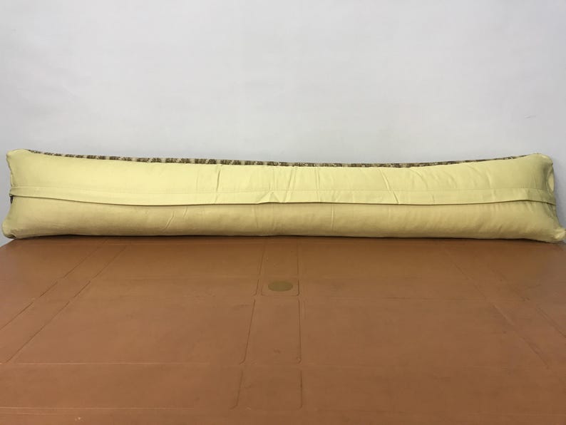 May include: A long, rectangular pillow with a light yellow back and a patterned top. The top features a mix of brown, green, and beige colors. The pillow is resting on a brown surface against a white background.