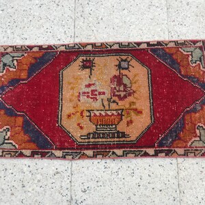 May include: Hand-woven rectangular rug with a central floral vase design. The rug has a red field with orange and blue borders. The central design is in shades of orange, yellow, and red. The rug has a fringed edge.