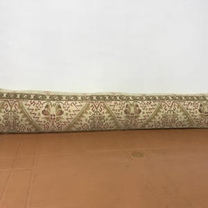 May include: A long, rectangular beige pillow with a detailed, repeating pattern in shades of brown, red, and green. The pattern includes geometric shapes and floral motifs. The pillow is resting on a brown surface.
