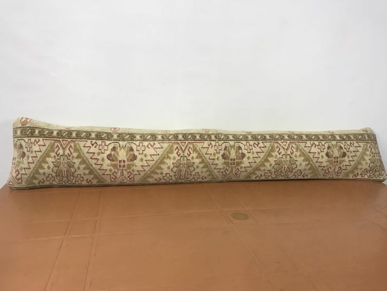 May include: A long, rectangular beige pillow with a detailed, repeating pattern in shades of brown, red, and green. The pattern includes geometric shapes and floral motifs. The pillow is resting on a brown surface.