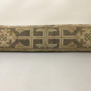 Peut inclure: Un chemin de table textile enroulé présentant un motif géométrique et floral répétitif dans des tons beige et gris sourd. Le tissu tissé affiche une surface texturée avec des motifs symétriques complexes disposés horizontalement.