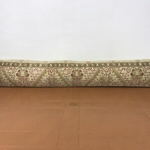May include: A long, rectangular pillow with a beige background and a repeating pattern in shades of brown and red. The pillow is placed on a brown surface.