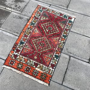 May include: A red, orange, and white patterned rug with a geometric design. The rug has a fringe border and is made of wool.