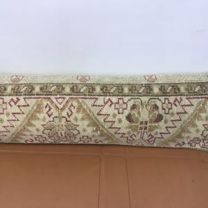 May include: Rectangular beige pillow with a detailed geometric pattern in shades of brown, red, and green. The pillow has a decorative border and is likely used for home decor.