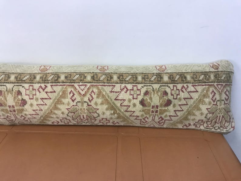 May include: Rectangular beige pillow with a detailed geometric pattern in shades of brown, red, and green. The pillow has a decorative border and is likely used for home decor.