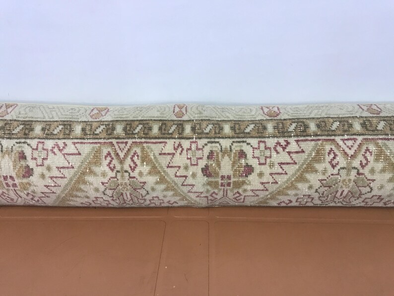 May include: Rolled-up beige and brown patterned rug. The rug features a repeating geometric design with floral elements in shades of beige, brown, and burgundy. The rug is rolled up, showing the detailed pattern.