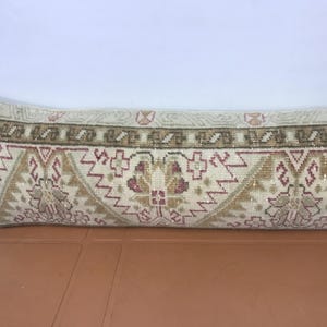 May include: A long, rectangular pillow with a patterned design. The pillow features a cream-colored background with a repeating pattern of geometric shapes in shades of brown, tan, and burgundy. The pillow is placed on a brown surface.