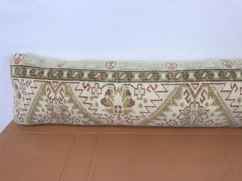 May include: A long, rectangular pillow with a patterned design. The pillow features a cream-colored background with a repeating pattern of geometric shapes in shades of brown, tan, and burgundy. The pillow is placed on a brown surface.
