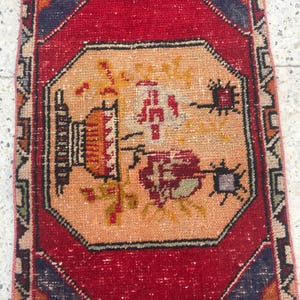 Puede incluir: Pequeña alfombra rectangular con un diseño octogonal central. La alfombra presenta un borde rojo y un campo central en tonos melocotón, amarillo y rojo, con patrones florales y geométricos. El diseño de la alfombra es simétrico y detallado.